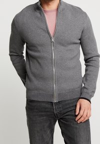 Grå zip-tröja med ståkrage, ribbade ärmslut och nederkant, tillverkad av ett mjukt material, bärs över en ljusrosa t-shirt.