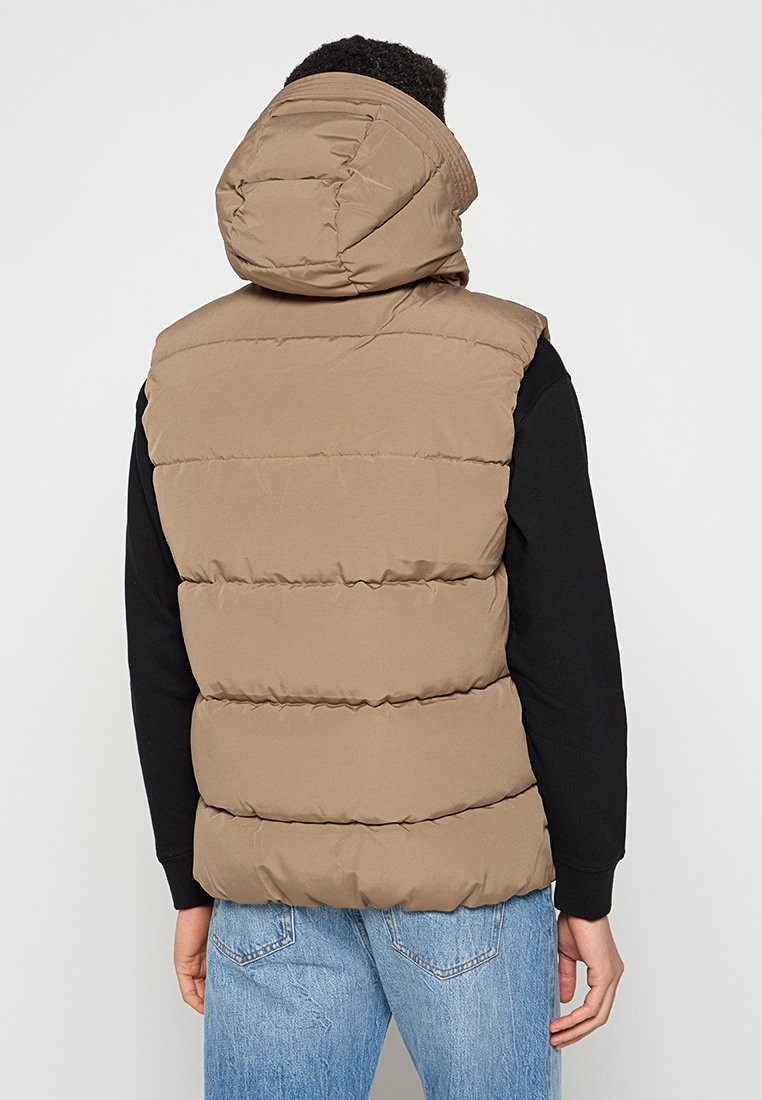 Beige Steppweste mit Kapuze, die horizontale gesteppten Abschnitte, eine glatte Textur aufweist und über einem schwarzen Langarmshirt getragen wird.