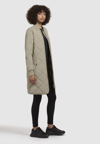 Cappotto imbottito e trapuntato di un beige chiaro con chiusura a zip e tasche laterali. Indossato sopra un top nero e abbinato a leggins neri e sneakers.