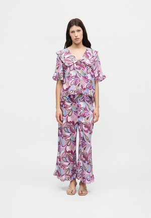 Jonge vrouw met lang donker haar die een bijpassende tweedelige outfit draagt met roze, paarse en blauwe wervelende patronen en ruches, staand op blote voeten.