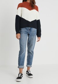 Sweter dzianinowy z wzorem color-block w kolorach rdzy, kremu i granatu, z okrągłym dekoltem i luźnym krojem. Połączony z podwiniętymi jeansami i czarnymi trampkami.