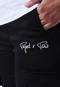 Pantaloni neri realizzati in morbido tessuto con un logo ricamato bianco che recita "Project x Paris" vicino alla tasca, con un design standard delle tasche.