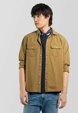 Giovane uomo con capelli scuri che indossa una camicia color sabbia con bottoni sopra una maglietta blu navy, jeans blu e una sciarpa al collo blu scuro, che guarda di lato.