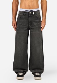 Suuria mustia farkkuhousuja korkealla vyötäröllä, joissa on valkoinen joustava vyötärönauha, jossa lukee "PEGADOR", sekä etutaskut. Rakenne on teksturoitu.