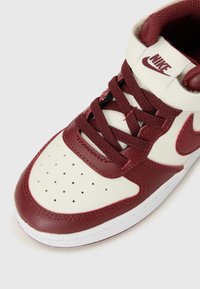 Červená a krémová Nike teniska s vysokým strihom, viditeľnými šnúrkami a perforovanou špičkou pre priedušnosť. Záber zblízka.