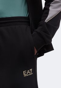 Άτομο που φοράει μαύρη φόρμα Emporio Armani EA7 με το χέρι στην τσέπη και μαύρο μπουφάν πάνω από μια τιρκουάζ μπλούζα.