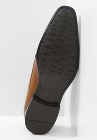 Chaussure en cuir marron avec une surface lisse, couture bleue le long de la semelle, et semelle extérieure en caoutchouc noir avec un détail de marquage en relief.
