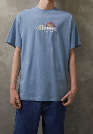 Person trägt ein hellblaues ellesse T-Shirt mit rotem und orangem Sonnenlogo, kombiniert mit dunkelblauen Jeans, vor einer grauen Betonwand.