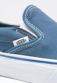 Blå slip-on sneakers i kanvas med vit söm, Vans-logotyp på etiketten och blå accentlinje på en vit gummisula. Närbild.