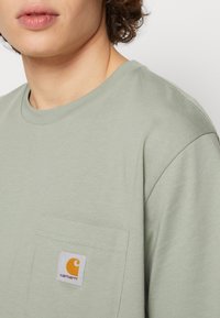 Lichtgroene katoenen t-shirt met een ronde hals en korte mouwen. Heeft een borstzak met een wit label en een oranje logo-detail.