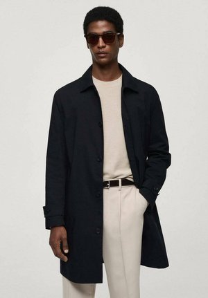 Cappotto nero in tessuto con chiusura a bottoni, indossato sopra un maglione beige chiaro. Tessuto liscio, vestibilità sartoriale, abbinato a pantaloni beige e occhiali da sole.
