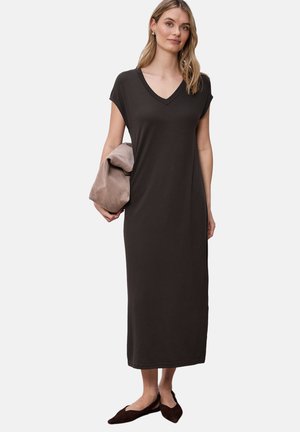 Vrouw die een mouwloze zwarte midi-jurk met V-hals draagt, een opgevouwen taupekleurig jasje vasthoudt en zwarte puntige ballerina's aan heeft, staand tegen een witte achtergrond.