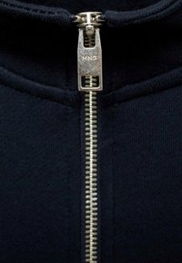 Giacca nera con zip frontale in metallo argentato. Il tessuto ha una texture liscia e un design aderente, con colletto alto.