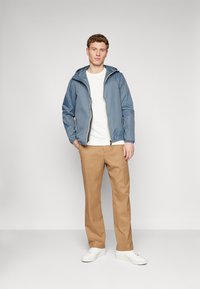 Lys blå vandtæt jakke med hætte, lynlåslukning og elastiske ærmer, parret med en hvid skjorte, beige bukser og hvide sneakers.