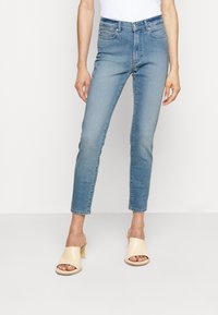Mujer vestida con unos pantalones vaqueros ajustados de color azul claro y sandalias de tacón abiertas de color beige, de pie contra un fondo blanco puro.