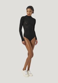 Svart långärmad bodysuit med en texturerad spetsmönster och hög krage, tillsammans med vita sneakers mot en ljus bakgrund.