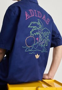 Mörkblå kortärmad skjorta med färgglad brodering av palmträd och strand. Har broderad "ADIDAS"-logotyp i rosa. Tillverkad i bomull.