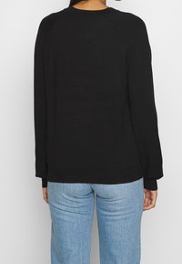 Zwarte sweater met lange mouwen, een ribbelhalslijn en manchetten, heeft een losse pasvorm en zachte, textuurgebreide stof. Draagt met lichtblauwe jeans.