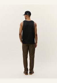 Black mesh tank top with white stripes, paired with brown checkered trousers and black shoes, viewed from the back. Cap worn on the head.