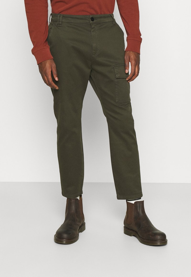 Olivgröna cargobyxor, slim fit, i bomullsmaterial, framfickor och en sidocargoficka. Bärs med bruna ankelstövlar.