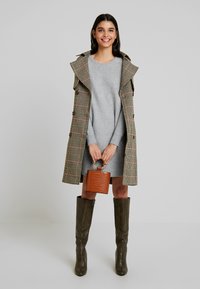 Ruda houndstooth modeli paltas, pilkas megztinis suknelė, alyvuogių spalvos kelio ilgio batai, laikantis mažą rudą tekstūruotą rankinę. Paprastas dizainas, struktūruotas siluetas.
