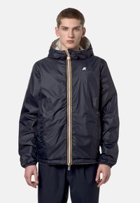 Jeune homme portant une veste à capuche bleu marine avec une fermeture éclair à l'avant et des poignets élastiques, debout devant un arrière-plan clair uni.