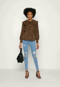 Blusa floral castanha com colarinho franzido, combinada com jeans azuis desgastados. A modelo segura uma mala preta e veste sandálias de salto pretas.