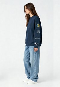 Felpa blu marina con grafiche che raffigurano un sole e scritte sulle maniche, abbinata a jeans baggy azzurri e sneakers bianche.