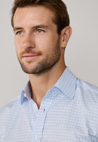 Man met lichtbruin haar en baard, gekleed in een lichtblauw geruit overhemd met knopen, kijkt enigszins opzij tegen een effen achtergrond.