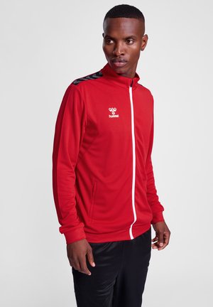Giacca sportiva rossa con zip bianca, colletto alto e dettagli neri sulle spalle. Realizzata in un materiale leggero e strutturato. Logo Hummel visibile.