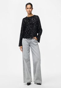 Mulher vestindo uma blusa de mangas longas pretas com lantejoulas, calças largas cinza claro, sapatos de salto pretos e brincos de argola prateados, de pé com a mão no bolso.