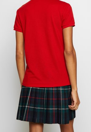 Personne portant une chemise rouge à manches courtes et une jupe plissée à carreaux vert foncé et bleu marine longueur genou, vue de dos.