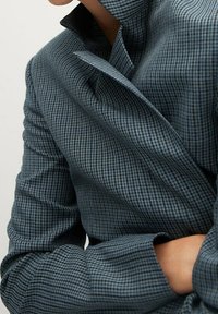 Veste à carreaux bleue et noire en tissu texturé, dotée d'un col montant et de manches longues avec des détails aux poignets.