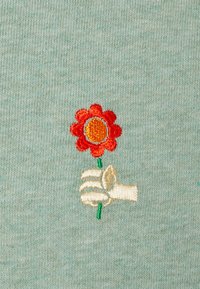 Vyšívaná nášivka zobrazující ruku držící červenou květinu se žlutým středem na texturovaném světle modro-zeleném látkovém pozadí.