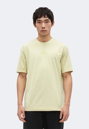 Ung mand iført en lysegul Adidas T-shirt og sorte bukser, stående mod en ensfarvet lys baggrund.