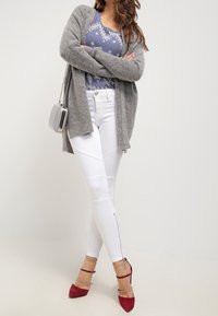 Šedý pletený kardigan, modré vzorované tričko, bílé upnuté džíny s kontrastním prošitím po stranách a červené lodičky na vysokém podpatku.