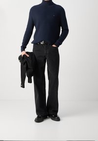 Personne portant un pull à col roulé bleu marine, ceinture noire et jean noir ample, tenant une veste noire dans la main gauche, debout sur un sol blanc.