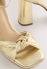 Zlaté sandále na vysokom opätku majú uzlový dizajn na nariasenom remienku, štvorcový tvar špičky a robustný kovový opätok. Hladká textura.