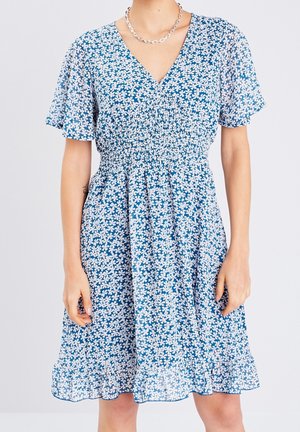 Robe bleue à manches courtes avec imprimé floral blanc, col en V, taille froncée et ourlet à volants, portée avec un collier chaîne argenté.