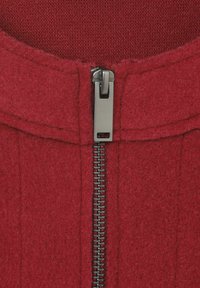 Close-up van een rode wollen jas met een metalen rits die deels is gesloten, waarbij de textuur van de stof en de stiksels zichtbaar zijn.