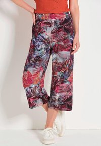 Frau trägt knöchellange, weit geschnittene Hosen mit bunten Blumen- und abstrakten Mustern, kombiniert mit weißen Sneakers und einem orangefarbenen Oberteil.