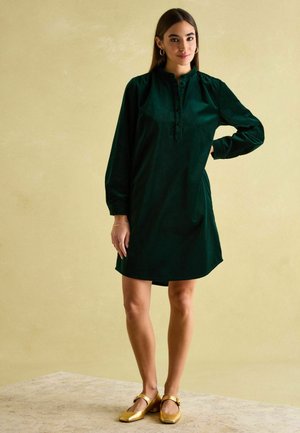 Dunkelgrünes, langärmliges Kleid mit einem knöpfbaren Ausschnitt, das eine lockere Passform und einen glatten Stoff aufweist, kombiniert mit goldenen Blockabsatzschuhen.