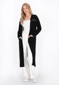Schwarzer, langer Cardigan mit weißen Paspeln, ausgestattet mit Taschen, aus weichem Stoff. Über einem weißen Oberteil getragen und kombiniert mit weiten weißen Hosen und Sneakers.