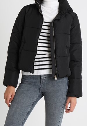 Personne portant une doudoune noire par-dessus un pull à col roulé blanc avec des rayures noires et un jean bleu clair.