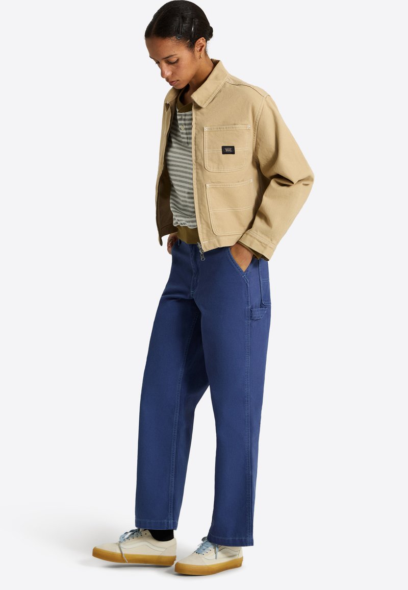 Femme portant une veste beige, une chemise rayée, un pantalon bleu à jambes larges et des baskets blanches, debout les mains dans les poches, regardant vers le bas.