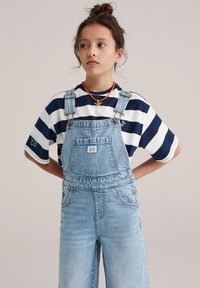 Salopetă din denim de culoare albastru deschis, cu accesorii reglabile argintii, buzunar frontal, combinată cu un tricou cu mâneci scurte în dungi alb și bleumarin.