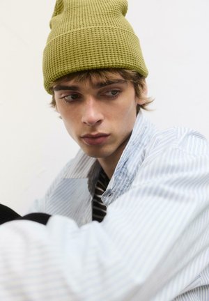 Zelený pletený čiapka, pruhovaná modrá košeľa s golierom a čierne detaily odevu; mäkké textúry, neutrálny pozadí.