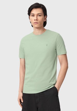 Ung mand med mørkt, glat hår iført en lysegrøn T-shirt med korte ærmer og sorte bukser, stående foran en ensfarvet baggrund.