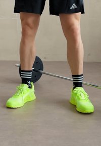 Neon green athletic shoes with textured mesh panels and rubber soles, paired with black striped socks and black shorts, beside a barbell.