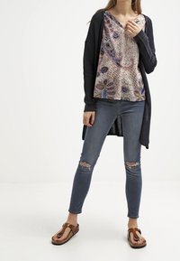 Largo cárdigan azul marino sobre una blusa de cuello en V con estampado en beige y acentos en azul, combinado con jeans claros desgastados y sandalias marrones.
